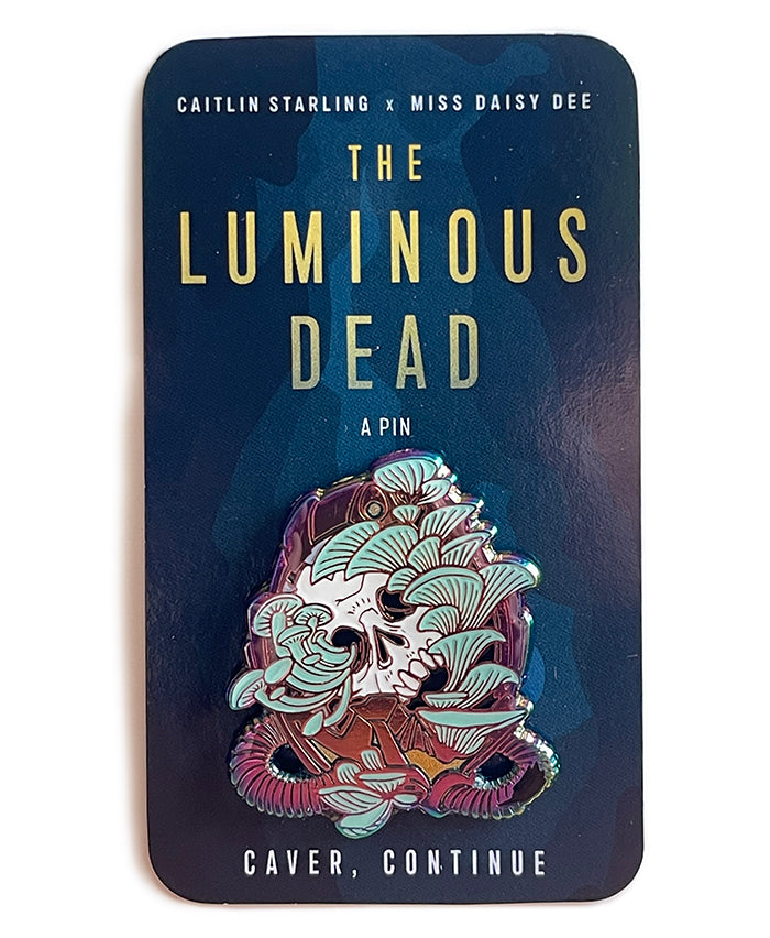 A Luminous Dead horror skull pin with pale teal blue mushrooms growing out of its orifices. The backing card is of a blue cave, and it says Caitlin Starling x Miss Daisy Dee, The Luminous Dead, a Pin, "Caver, continue." On a white background.